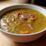 Close-up of a white bowl filled with thick, yellow pea soup topped with diced ham and carrots.