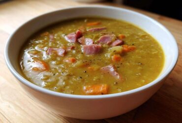 Close-up of a white bowl filled with thick, yellow pea soup topped with diced ham and carrots.