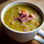Close-up of a white mug filled with thick, green pea soup topped with chunks of ham and carrots.