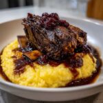 A close-up of tender braised short ribs served atop a bed of creamy yellow polenta, drizzled with rich sauce and fruit topping.