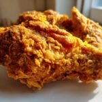 Close-up shot of a piece of golden brown, extra crispy fried chicken resting on a white plate.