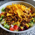A close-up view of a hearty taco salad featuring seasoned ground beef, lettuce, beans, corn, and topped with cheese sauce and corn chips.