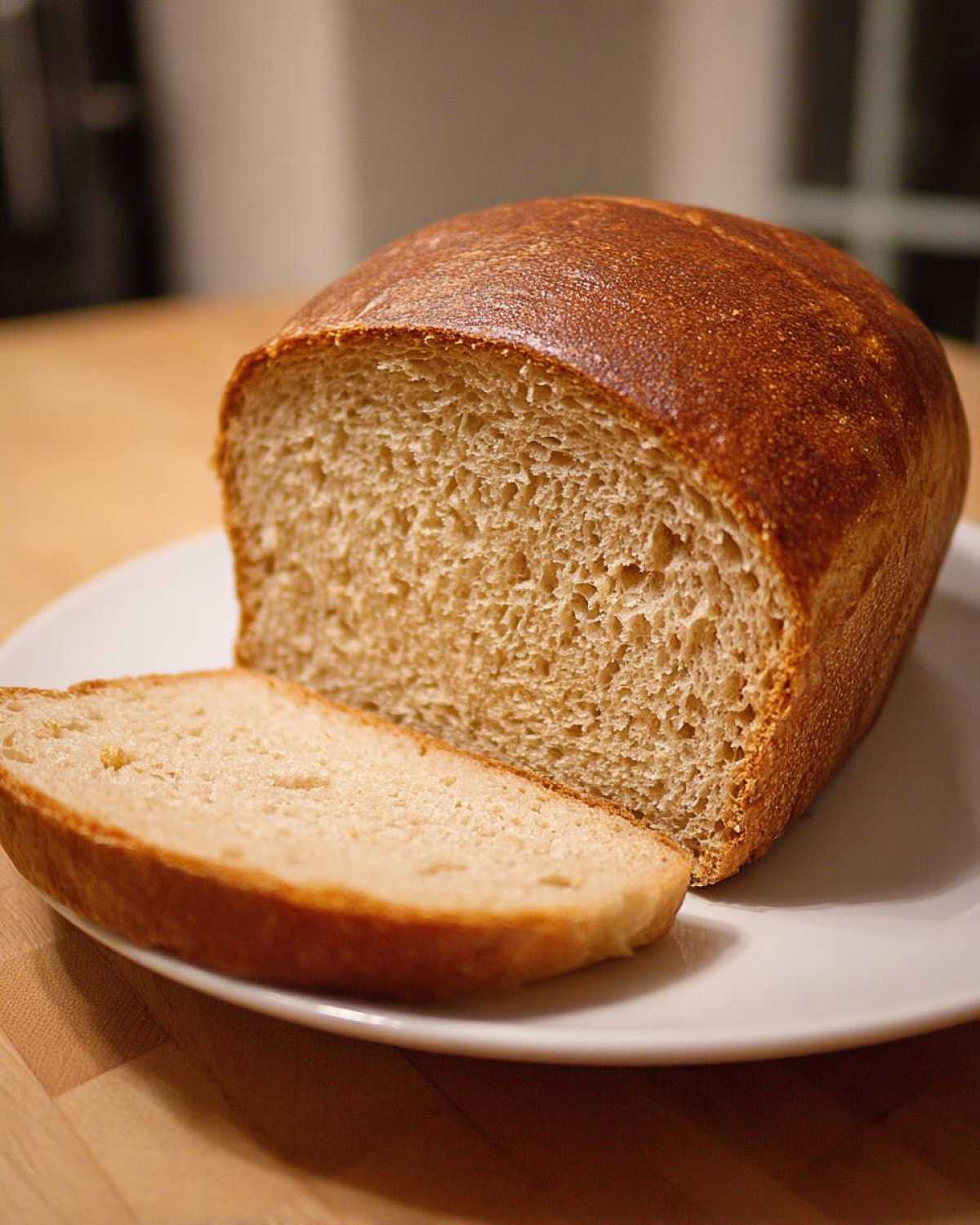 A freshly baked loaf of whole wheat bread, partially sliced, resting on a white plate.