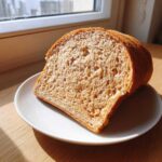 A perfect slice of homemade whole wheat bread showing its airy texture, resting on a white plate near a window.