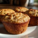 Close-up of four golden-brown banana protein muffins resting on a white plate, highlighted by natural sunlight.