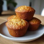 Four golden-brown banana protein muffins stacked on a light grey plate, ready to eat.