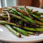 A close-up of blistered chinese green beans with garlic sauce piled on a white plate.