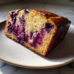 Close-up of a moist slice of blueberry banana bread showing vibrant purple blueberry swirls in the golden crumb.