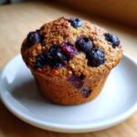 A single, golden-brown blueberry protein muffin topped with baked blueberries, resting on a small white plate.