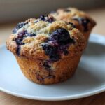 A close-up of a freshly baked blueberry protein muffin with a crumbly top, sitting on a white plate.