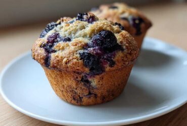 A close-up of a freshly baked blueberry protein muffin with a crumbly top, sitting on a white plate.