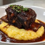 A single, rich braised short ribs portion served atop creamy yellow polenta, drizzled with dark sauce and garnished with thyme.