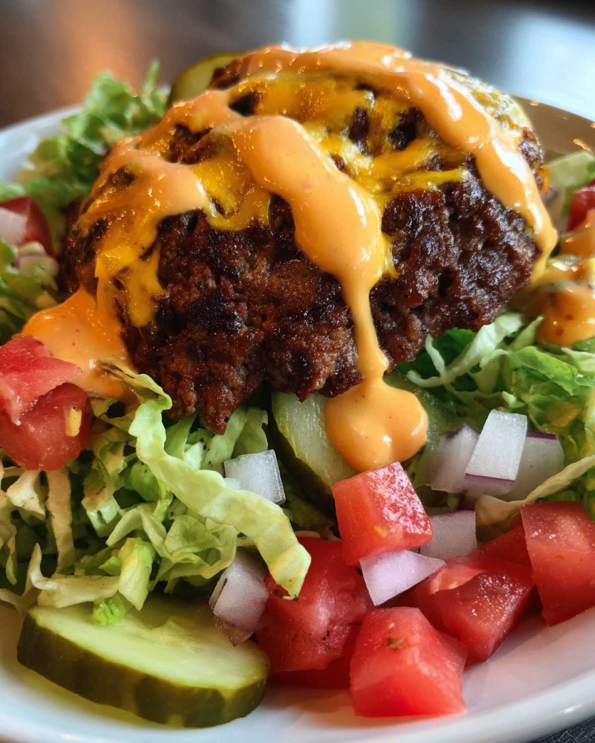 A close-up of a juicy cheeseburger bowl featuring a seasoned patty topped with melted cheese and special sauce over lettuce, pickles, tomatoes, and onions.