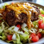 Close-up of a juicy beef patty topped with melted cheese sauce in a fresh cheeseburger bowl over lettuce, tomatoes, and onions.