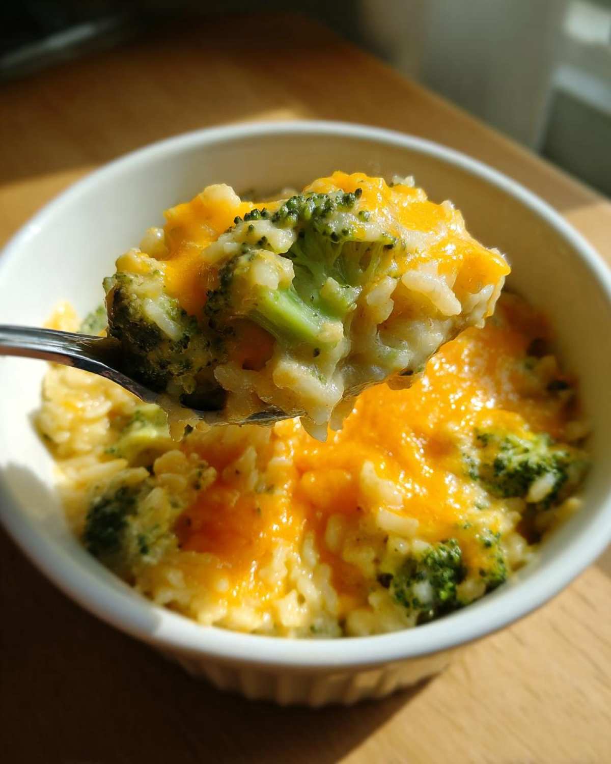 A spoonful of creamy, cheesy broccoli rice lifted from a white bowl, showing melted cheddar cheese and bright green broccoli florets.