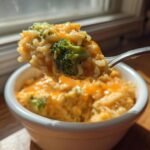 A fork lifts a scoop of creamy, cheesy broccoli rice out of a white bowl, backlit by window light.