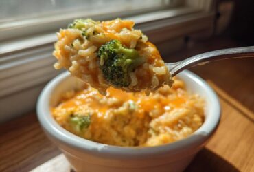 A fork lifts a scoop of creamy, cheesy broccoli rice out of a white bowl, backlit by window light.