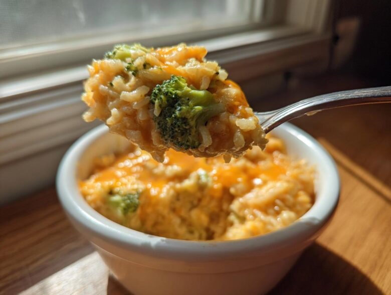 A fork lifts a scoop of creamy, cheesy broccoli rice out of a white bowl, backlit by window light.