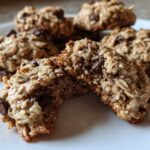 A chocolate chip oatmeal cookie broken in half, showing its chewy interior texture on a white plate.
