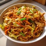 A close-up of a white bowl filled with homemade chicken chow mein recipe, featuring noodles, chicken pieces, cabbage, and carrots.