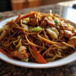 Close-up of a heaping plate of savory chicken chow mein recipe with noodles, chicken pieces, carrots, and cabbage.