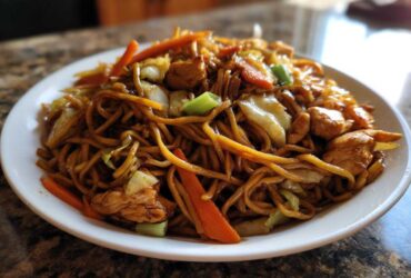 Close-up of a heaping plate of savory chicken chow mein recipe with noodles, chicken pieces, carrots, and cabbage.