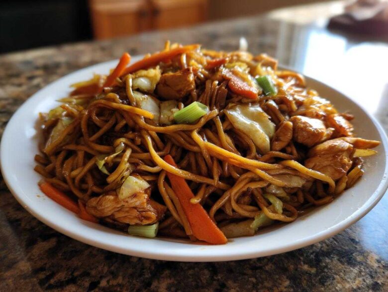 Close-up of a heaping plate of savory chicken chow mein recipe with noodles, chicken pieces, carrots, and cabbage.