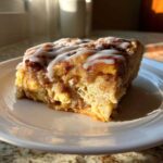 A close-up shot of a single serving slice of warm Cinnamon roll casserole topped with white icing.