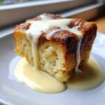 A close-up of a square slice of golden brown bread pudding topped with rich vanilla custard sauce.
