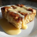 A square slice of classic bread pudding, golden brown and topped with rich vanilla sauce.