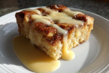 A square slice of classic bread pudding, golden brown and topped with rich vanilla sauce.