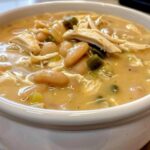 A close-up view of a white bowl filled with creamy crockpot white chicken chili, showing shredded chicken, white beans, and green chilies.