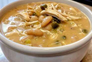 A close-up view of a white bowl filled with creamy crockpot white chicken chili, showing shredded chicken, white beans, and green chilies.