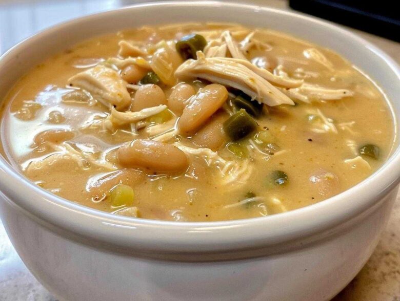 A close-up view of a white bowl filled with creamy crockpot white chicken chili, showing shredded chicken, white beans, and green chilies.