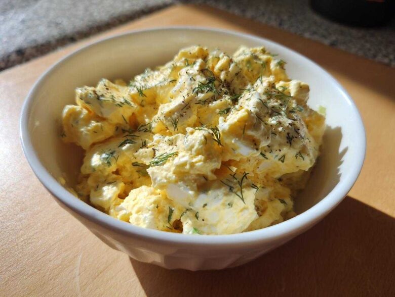 Close-up of a creamy egg salad recipe topped with fresh dill and black pepper in a white bowl.