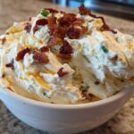A close-up of creamy Million Dollar Dip topped generously with crispy bacon bits and chives in a white bowl.