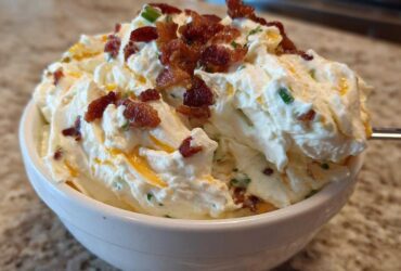 A close-up of creamy Million Dollar Dip topped generously with crispy bacon bits and chives in a white bowl.