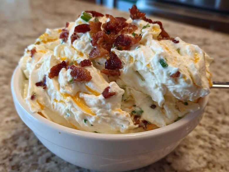 A close-up of creamy Million Dollar Dip topped generously with crispy bacon bits and chives in a white bowl.