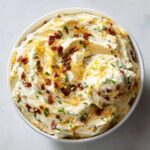 Overhead view of a bowl filled with creamy Million Dollar Dip, topped generously with shredded cheddar cheese, bacon bits, and fresh chives.