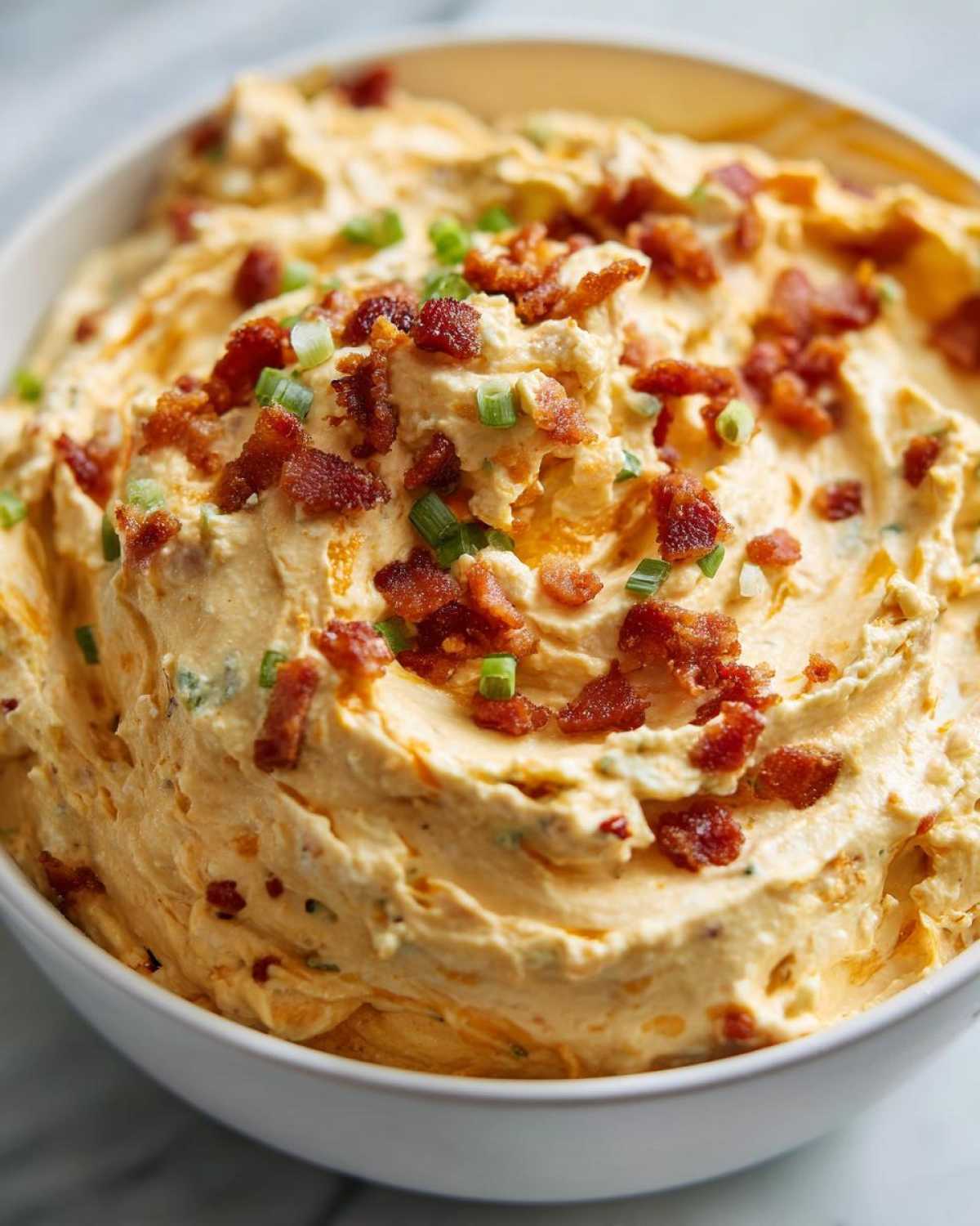 Close-up of creamy, orange-hued Million Dollar Dip topped generously with crumbled bacon and chopped green onions.