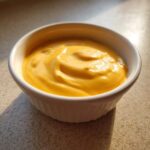 A small white ramekin filled with thick, bright orange, creamy nacho cheese sauce.