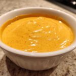 Close-up of vibrant orange, creamy nacho cheese sauce speckled with red spices in a small white bowl.