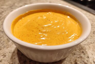 Close-up of vibrant orange, creamy nacho cheese sauce speckled with red spices in a small white bowl.