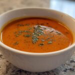 A close-up of a white bowl filled with rich, orange-red tomato bisque, topped with chopped green herbs.