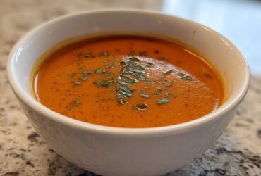A close-up of a white bowl filled with rich, orange-red tomato bisque, topped with chopped green herbs.