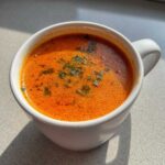 Close-up of a steaming, creamy tomato bisque in a white mug, topped with fresh green herbs.