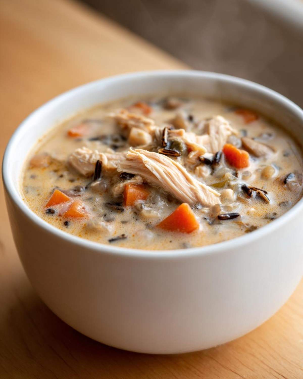Close-up of a steaming white bowl filled with creamy wild rice soup, featuring shredded chicken and bright orange carrots.