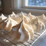 Close-up of several perfectly piped, light-colored meringue cookies cooling on a wire rack near a window.
