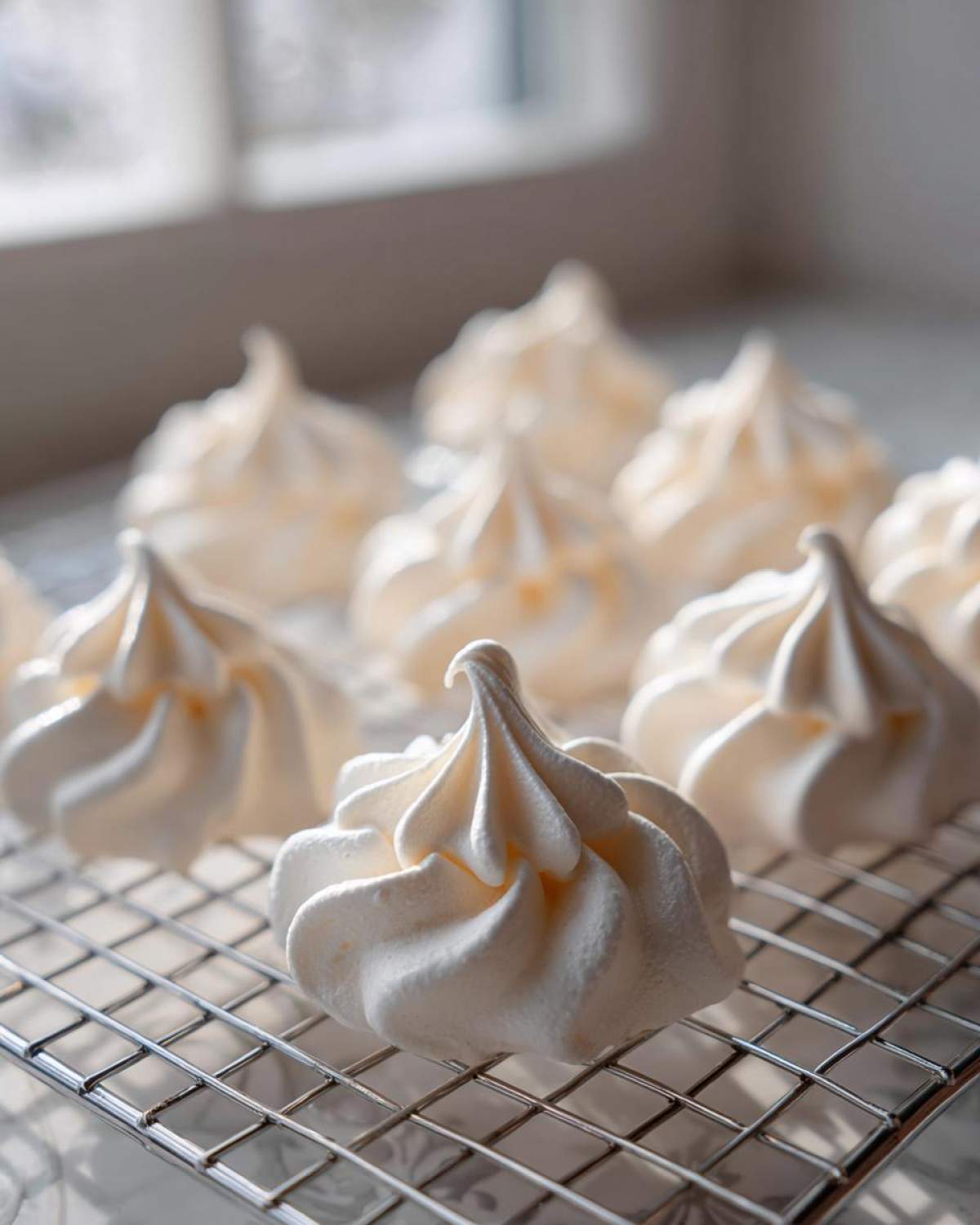 Close-up of freshly piped, white meringue cookies cooling on a wire rack near a bright window.