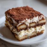 Close-up of a layered slice of tiramisu recipe showing ladyfingers, mascarpone cream, and cocoa powder dusting.